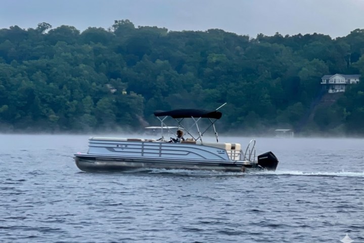 Pontoon boat with canopy on a lake, trees and house in the background.