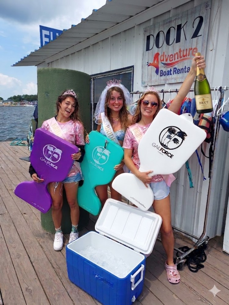 Three women in tiaras hold swim boards and a champagne bottle at a boat rental dock.