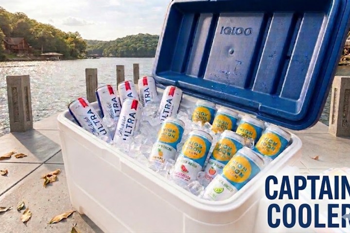 Open cooler by lake with canned drinks inside, labeled 'Captain Cooler'.