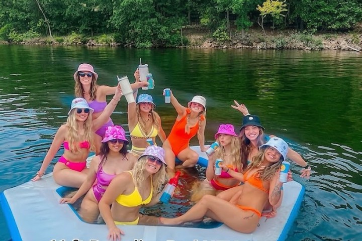 Group of women in swimsuits on a floating pad in a lake, surrounded by trees.