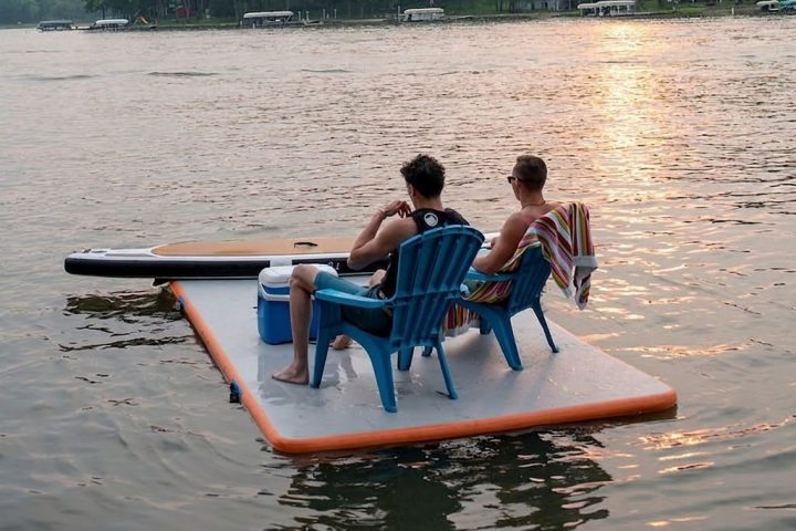 Two people sit on a floating pad with a cooler and chair on a calm lake at sunset.