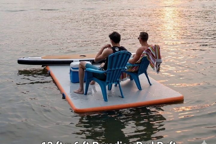 Two people sit on a floating pad with a cooler and chair on a calm lake at sunset.