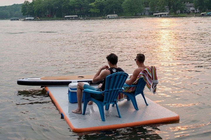 Two people sit on a floating pad with a cooler and chair on a calm lake at sunset.