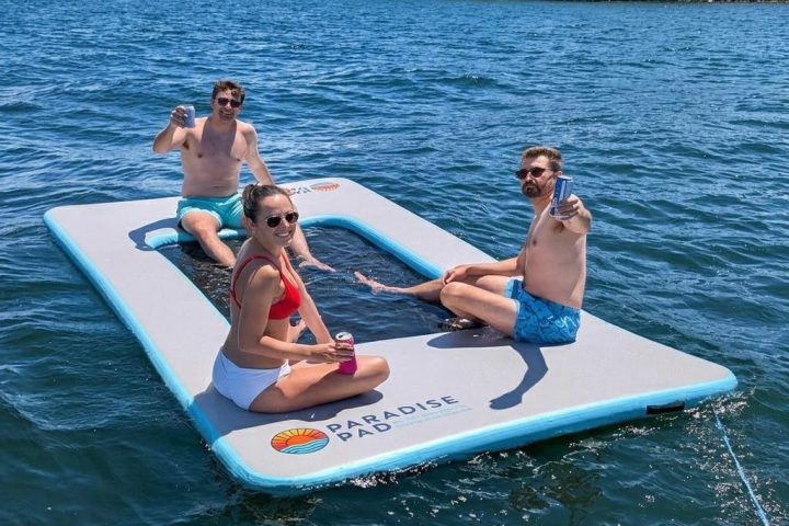 Three people relaxing on a floating pad in a lake, holding drinks, on a sunny day.