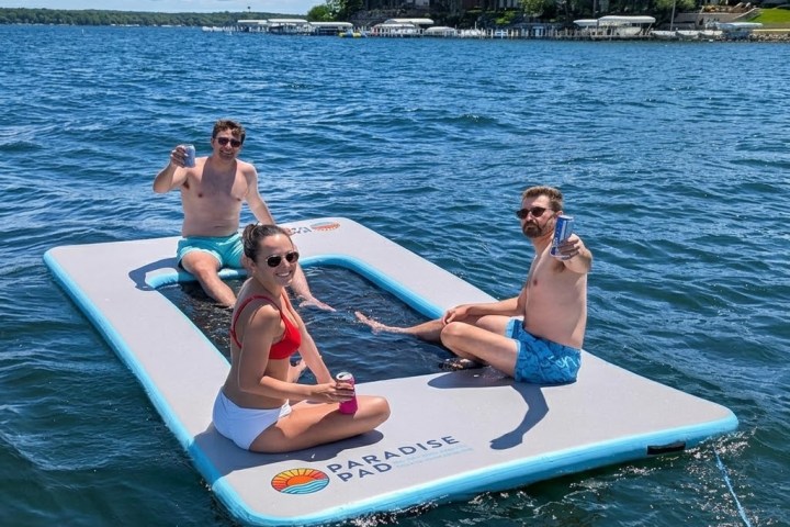 Three people relaxing on a floating pad in a lake, holding drinks, on a sunny day.