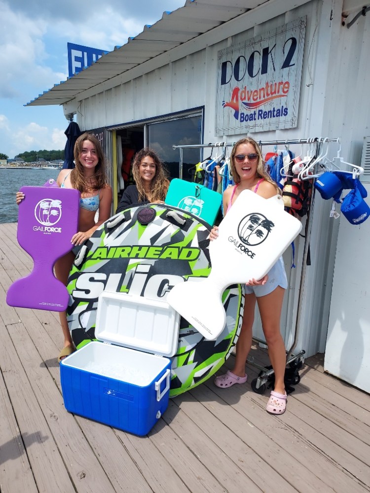 Three women holding water sports gear and a cooler on a dock by a boat rental shop.