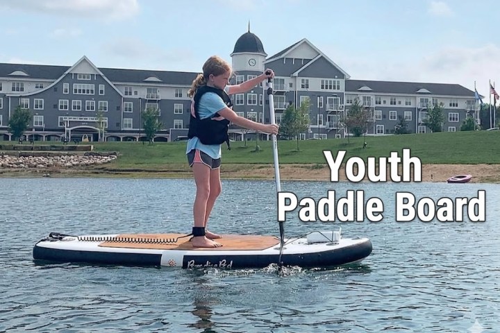 Child paddle boarding on a lake near a large building, text reads 'Youth Paddle Board.'